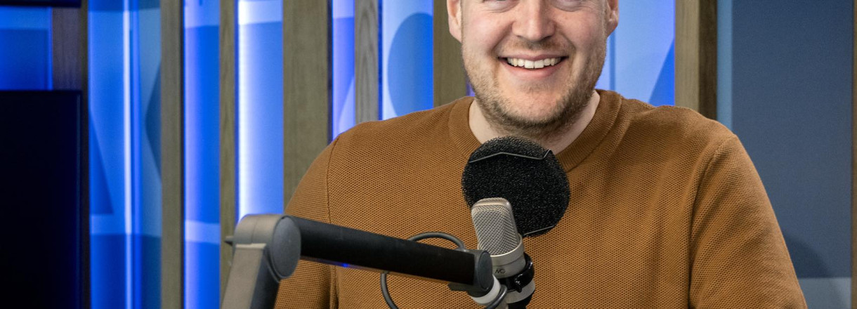 Bilde av Fredrik Johan Helland i eit radiostudio. Han står bak ein mikrofon og smiler.