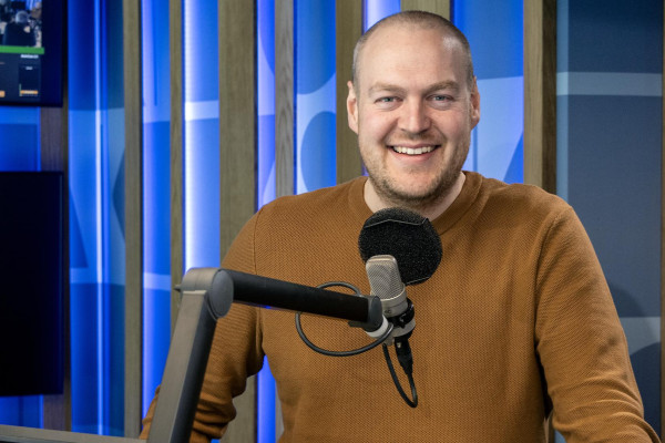 Bilde av Fredrik Johan Helland i eit radiostudio. Han står bak ein mikrofon og smiler.