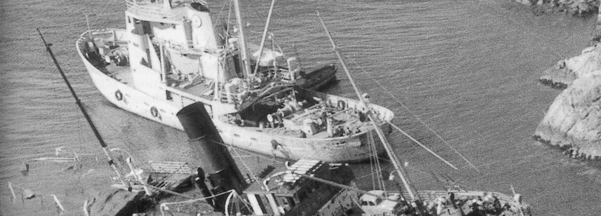 Svart kvitt bilete av to gamle båtar, der den eine ligg delevis ned i vatnet etter eit forlis. Det er ved ei kai og folk står på land og ser på båtane.